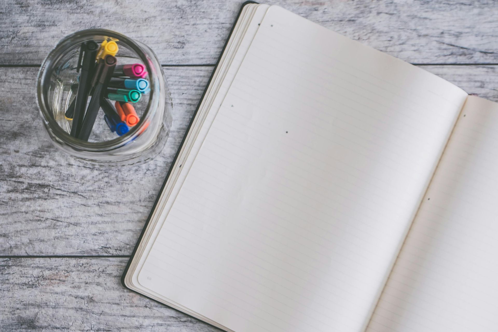Open notebook with blank pages and a glass jar of colorful pens on a wooden surface.