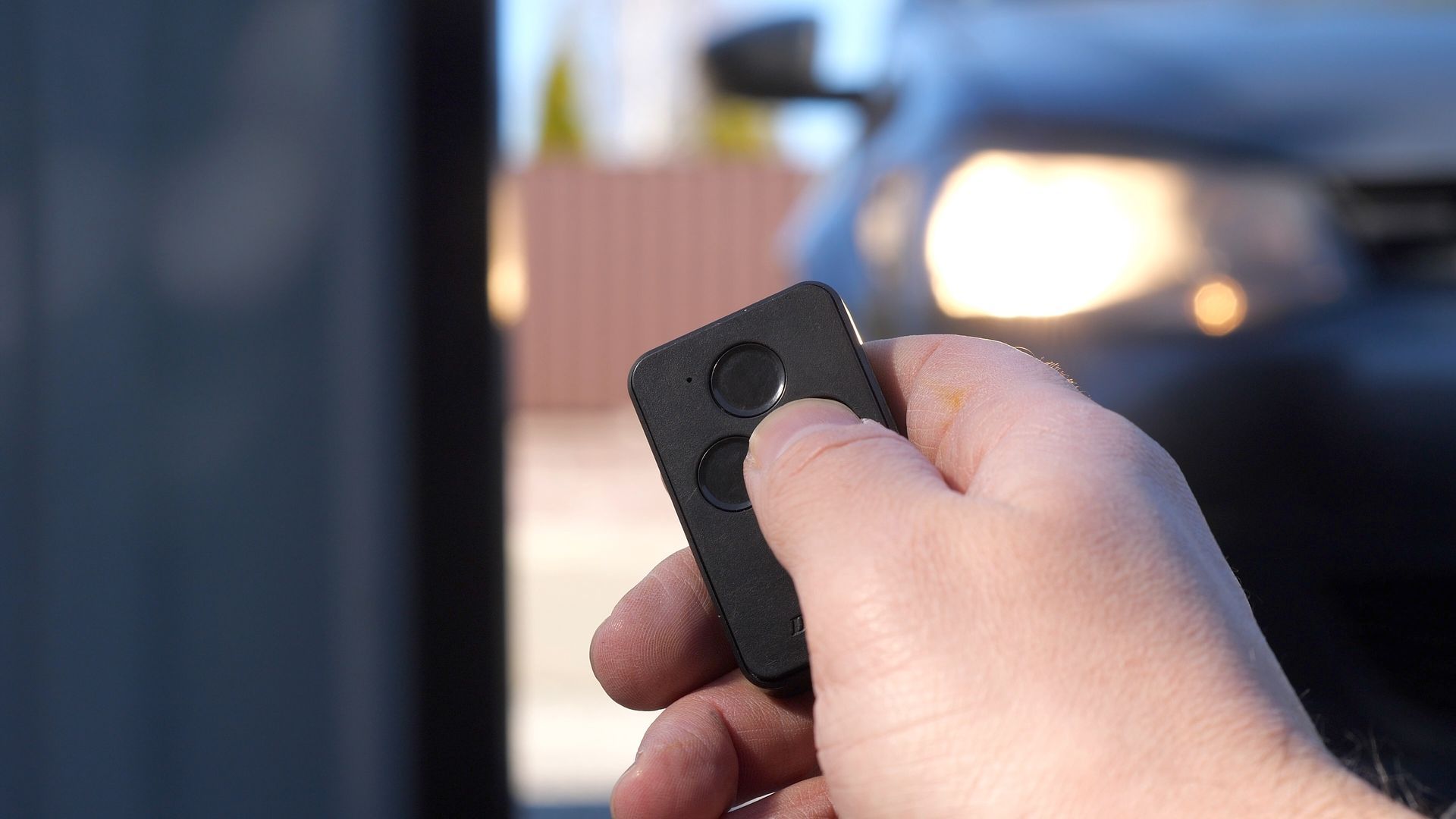A person holding a remote control with two buttons, pointing it toward a car with its headlights on.