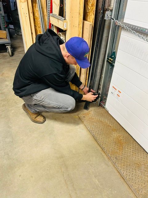 A person in a blue shirt repairs a garage door mechanism, attaching a cable to a bracket near the metal track. A person in a blue shirt repairs a garage door mechanism, attaching a cable to a bracket near the metal track.