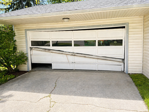 A hand holds a remote control, operating a white paneled garage door as it rises in front of a stone-faced building. A hand holds a remote control, operating a white paneled garage door as it rises in front of a stone-faced building.