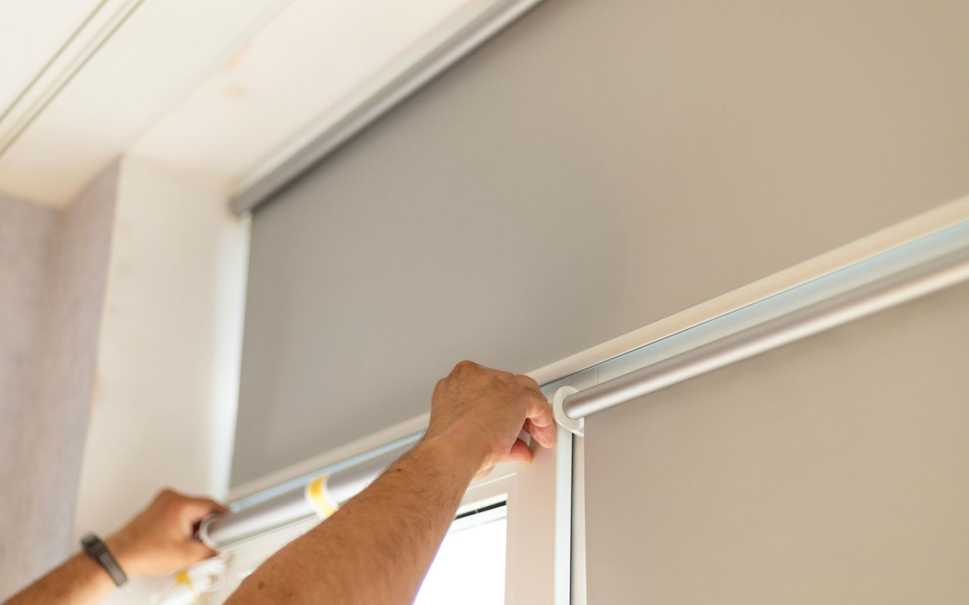 Hands adjusting a gray roller blind mounted on a window frame.
