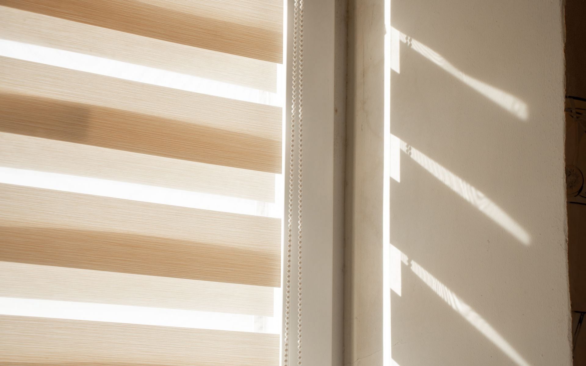 Beige and white striped window blinds filter sunlight, casting diagonal shadows onto a nearby white wall.