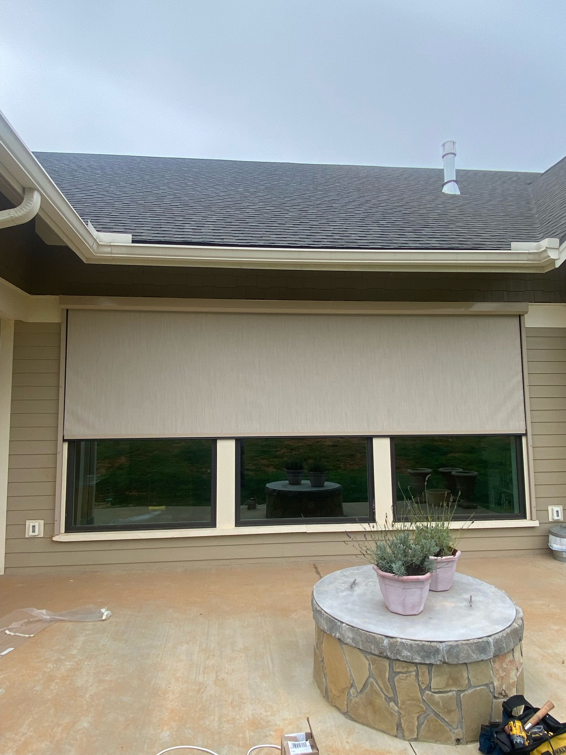 House exterior with closed beige shade, dark shingled roof, and a round stone planter on the patio