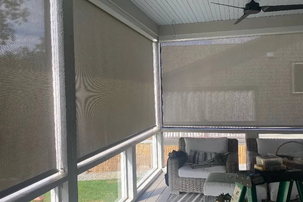 A screened-in porch with light-colored roller shades partially lowered over windows, featuring patio furniture inside.