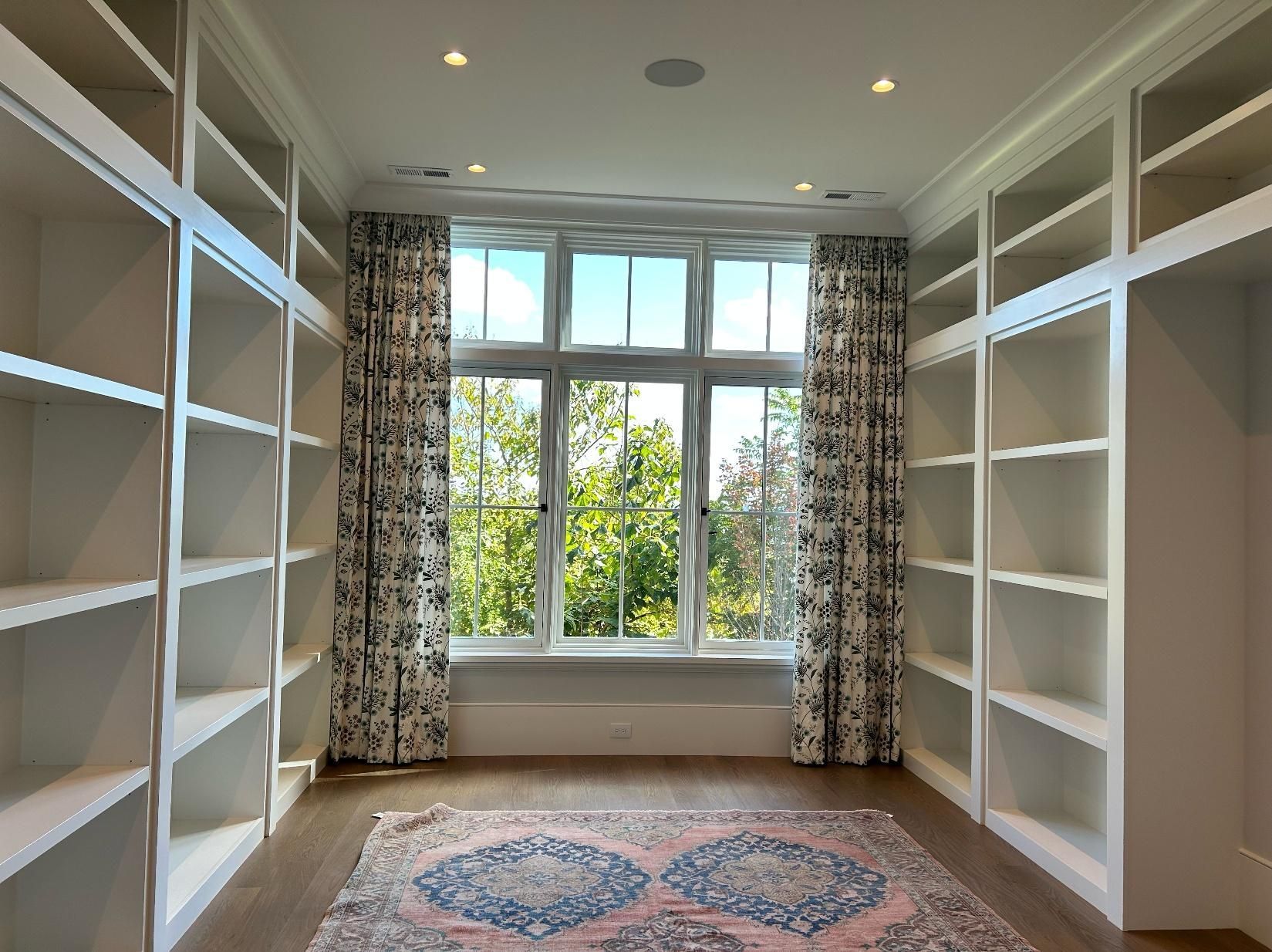Empty walk-in closet with white built-in shelves, patterned curtains, and a window over a round rug