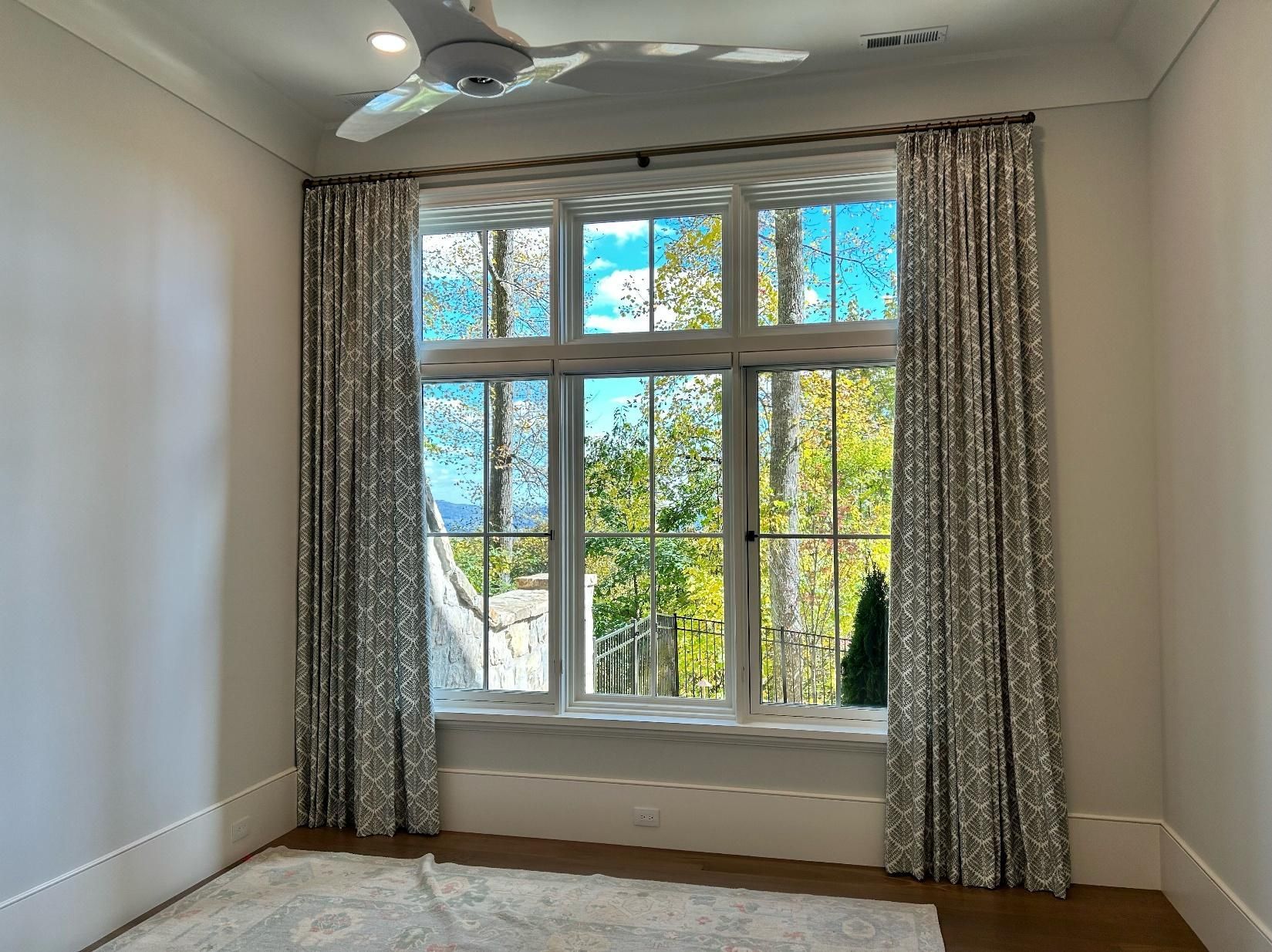 Empty room with white walls, beige carpet, ceiling fan, and large windows with curtains and greenery outside
