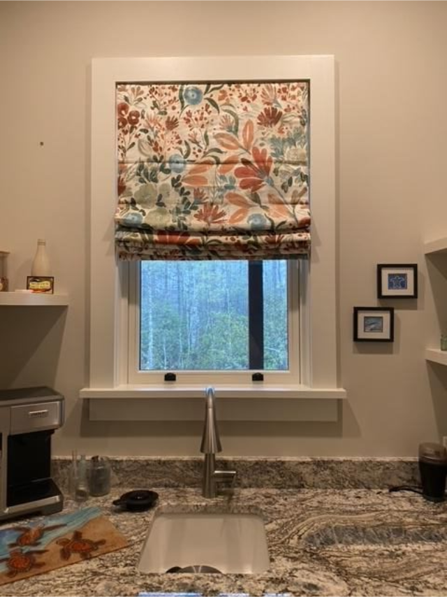 Bathroom sink beneath floral Roman shade over a window, with granite counter and wall shelves.