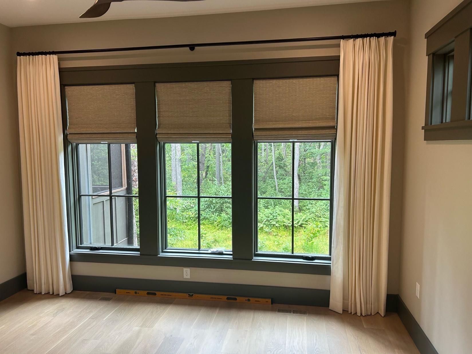 Empty living room with large window and cream curtains, overlooking greenery outside.