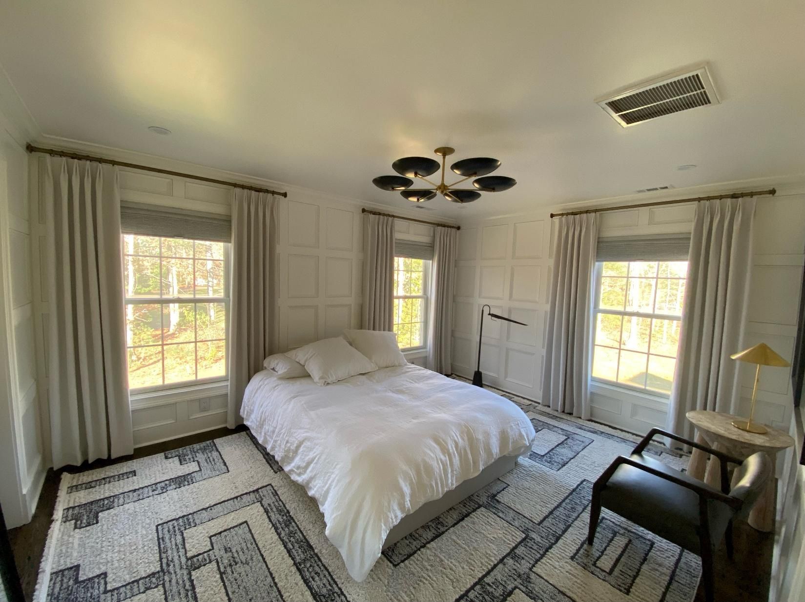 Bright bedroom with a white bed, patterned rugs, tall windows, and a small side table with lamp