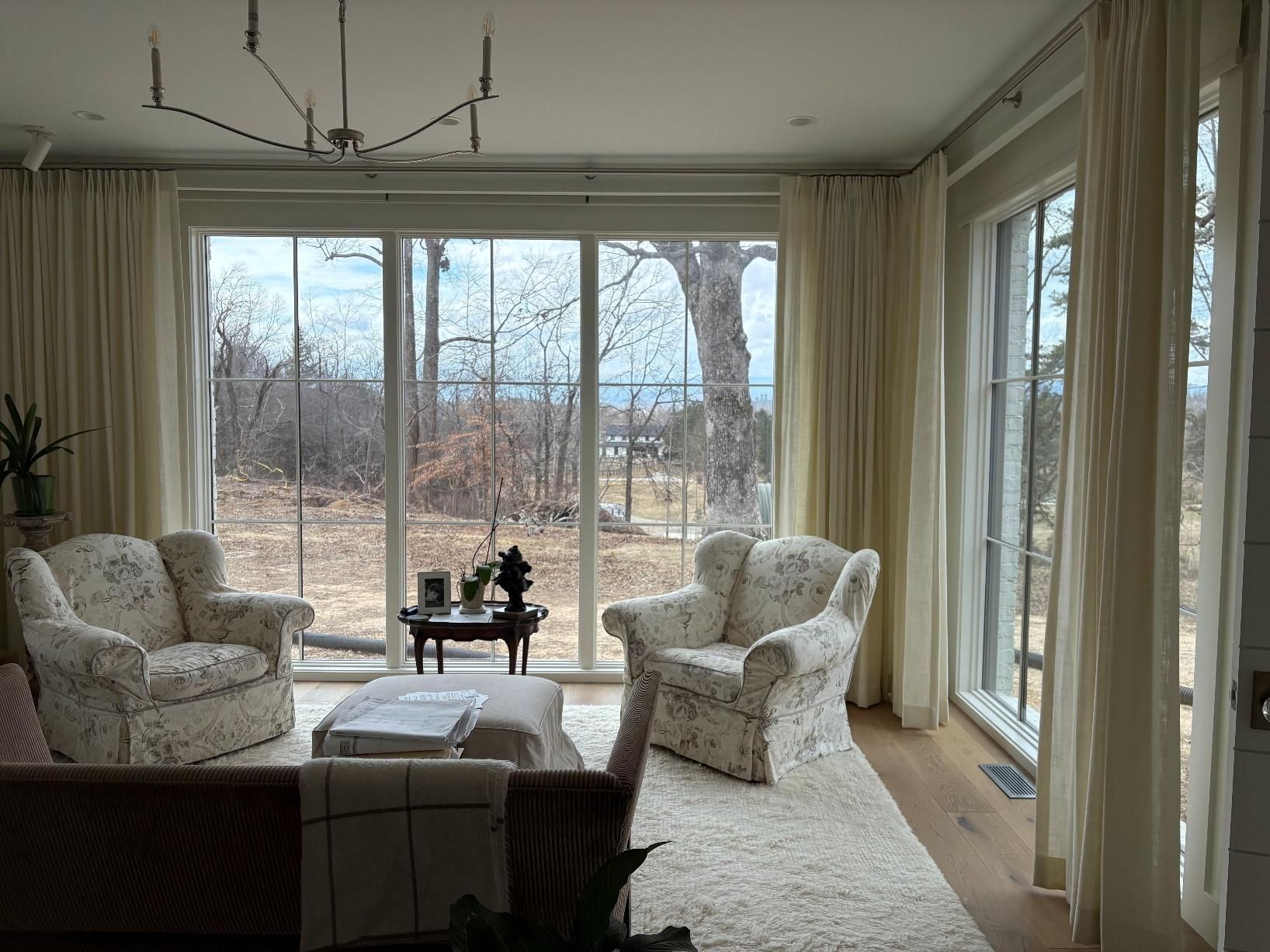 Bright living room with white armchairs, a sofa, and floor-to-ceiling windows overlooking a wooded yard