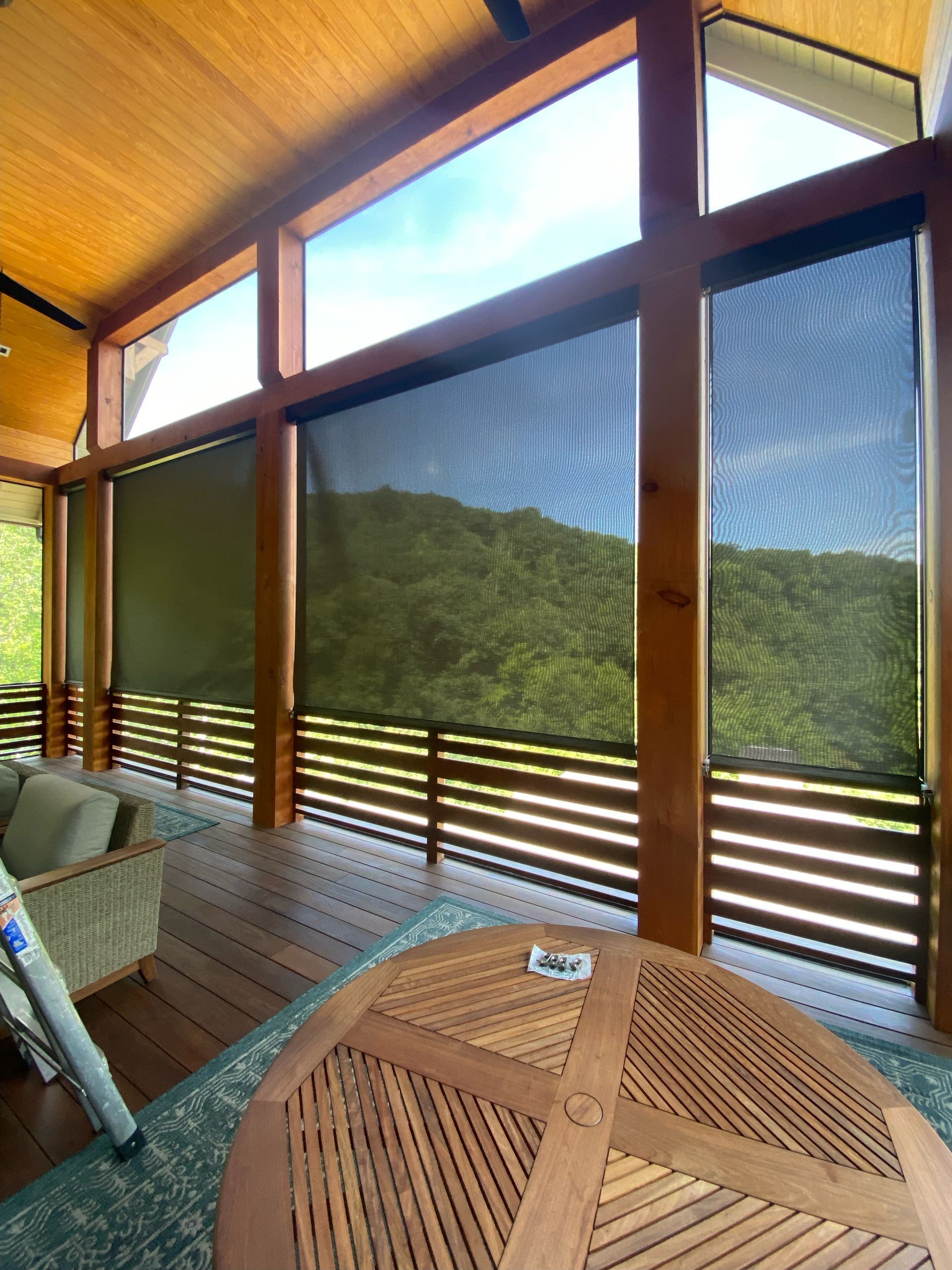 Wooden porch with woven chairs and round table, overlooking green hills through large screened windows.