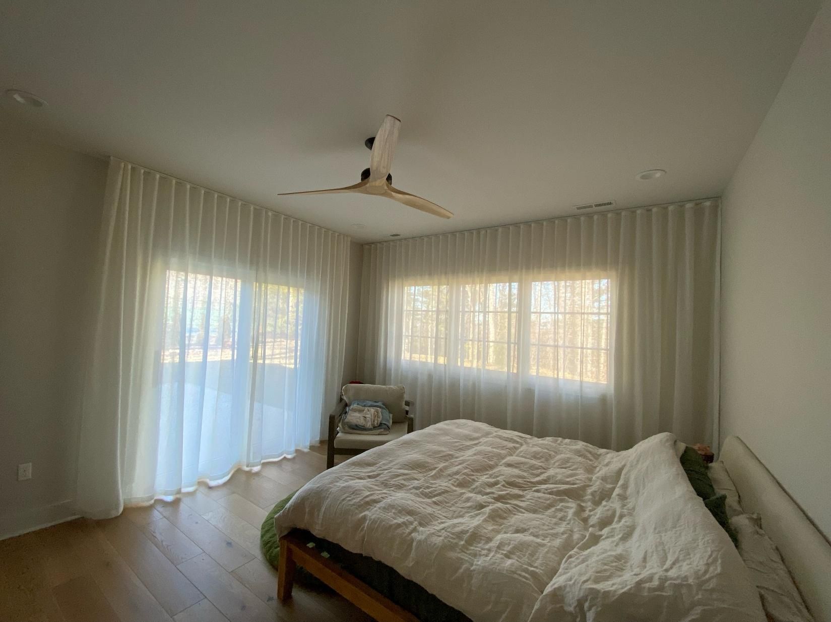 Bright bedroom with sheer curtains, large windows, a ceiling fan, and an unmade bed near a chair.