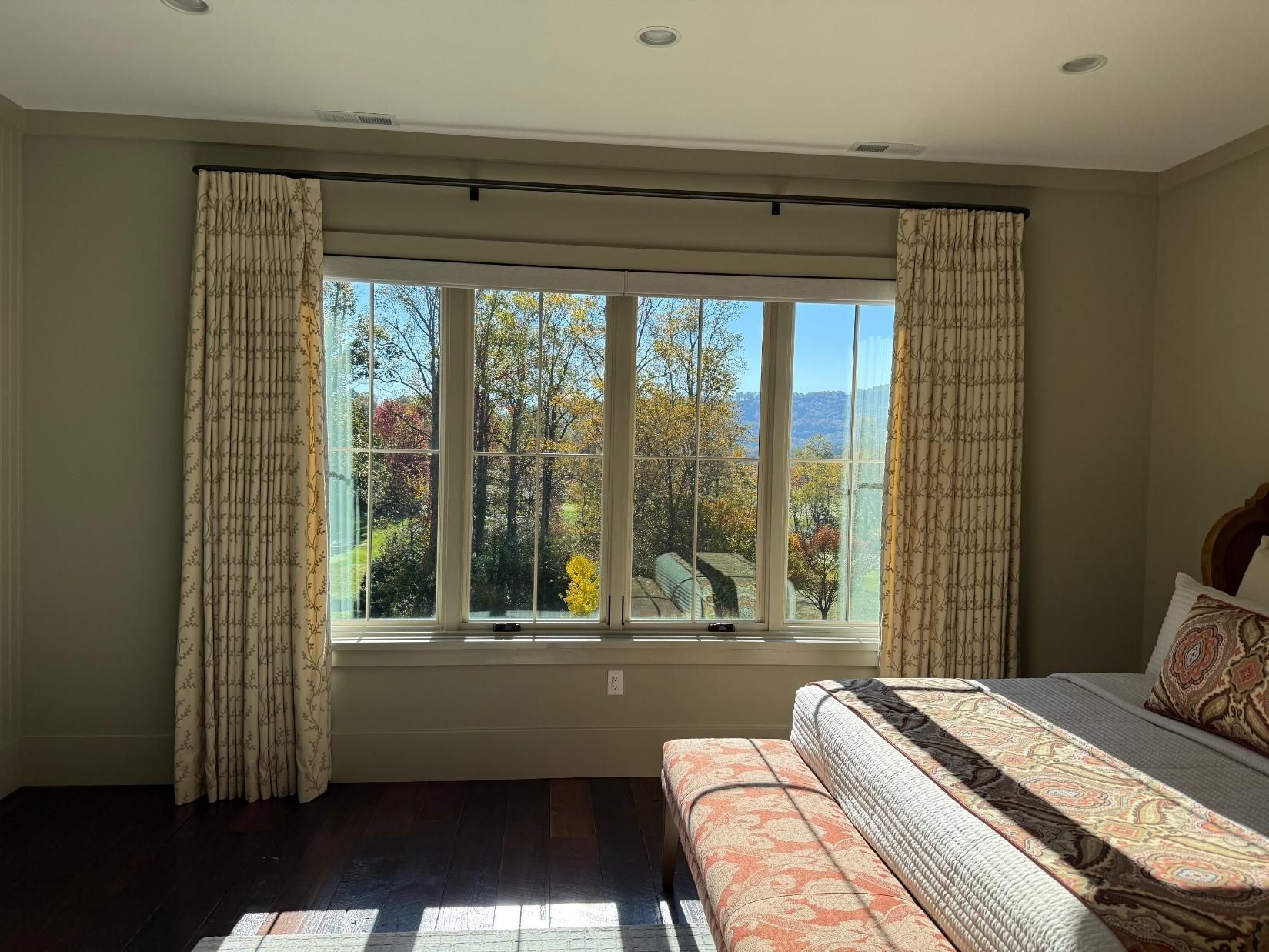Sunlit bedroom with large window, patterned curtains, and a bed with quilted bedding.
