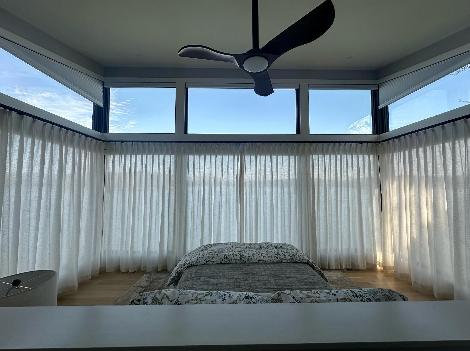 Bright bedroom with a bed, sheer curtains, large windows, and a ceiling fan under a blue sky.