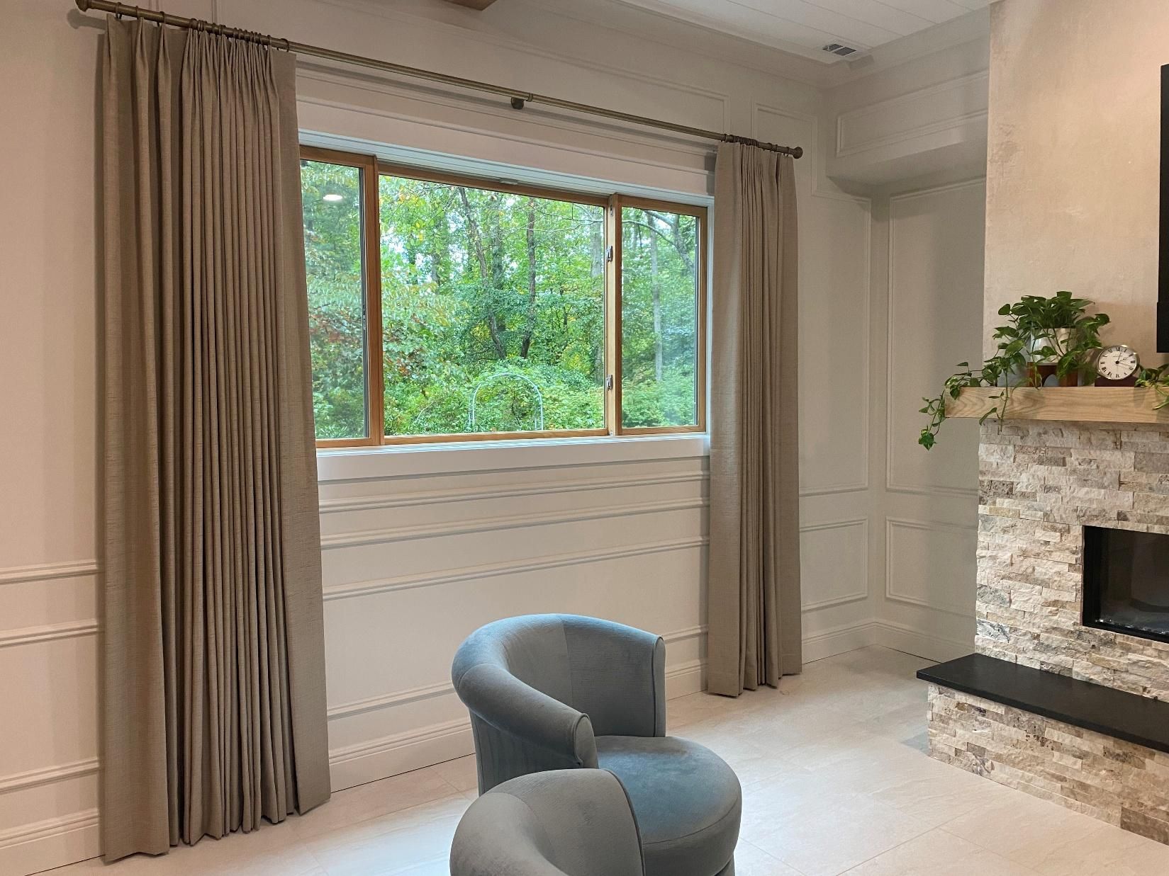 Bright living room with gray chair, large window, beige curtains, and stone fireplace.
