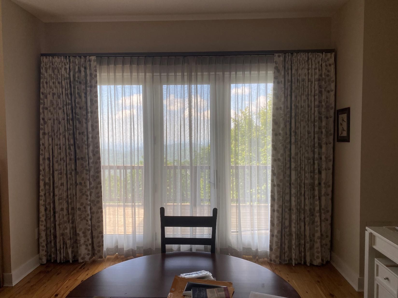 Dining room with a round table and chair facing large windows with sheer curtains and patterned drapes.