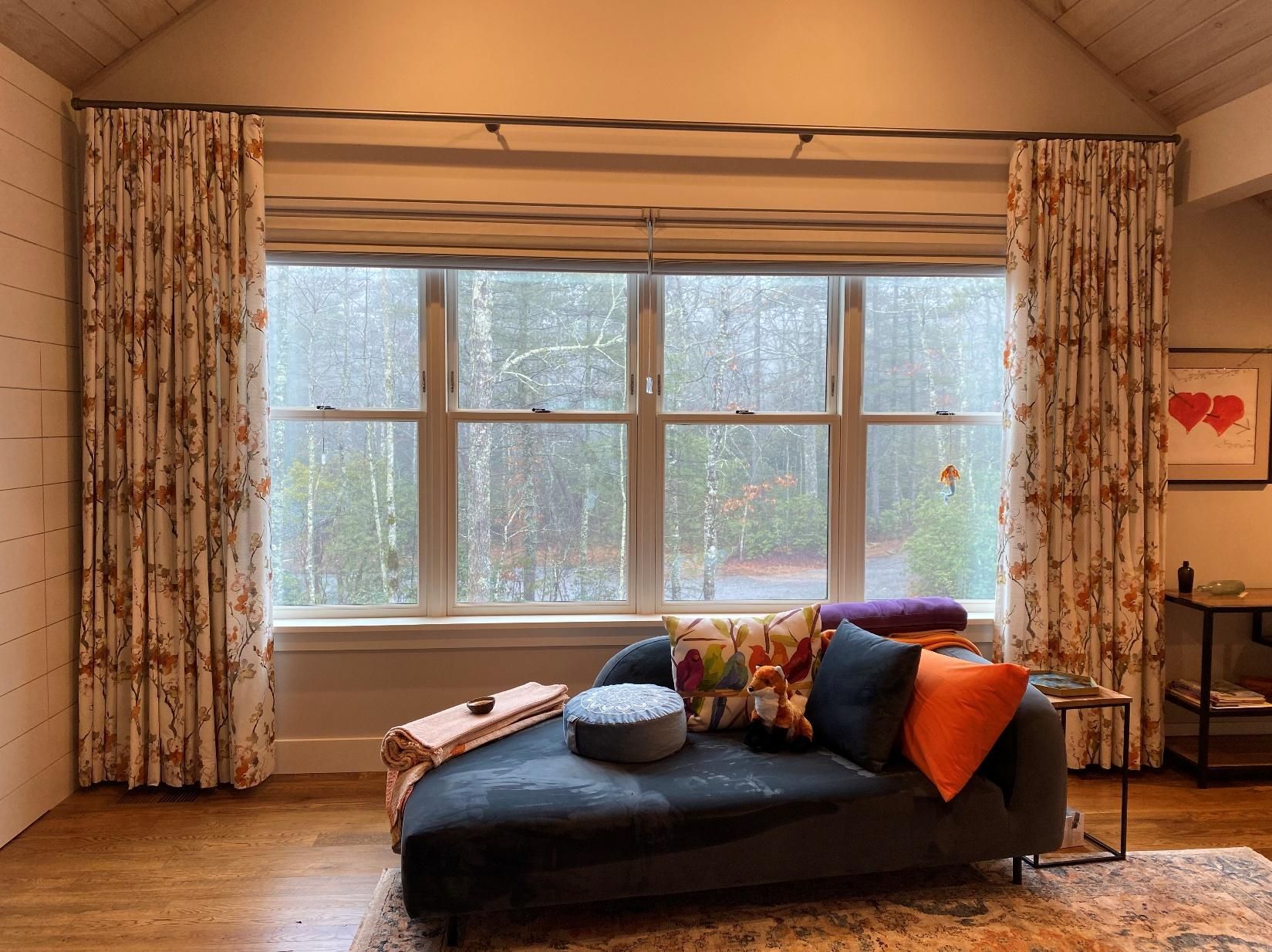 Living room with large rain-streaked window, floral curtains, and a dark chaise lounge with colorful pillows.