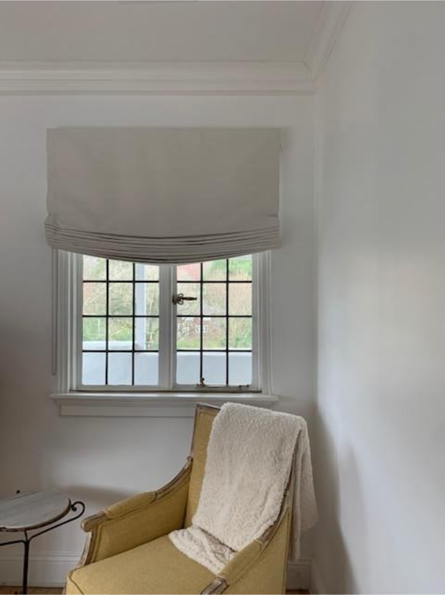 Cozy corner with a cream chair, draped blanket, small table, and window with a Roman shade.