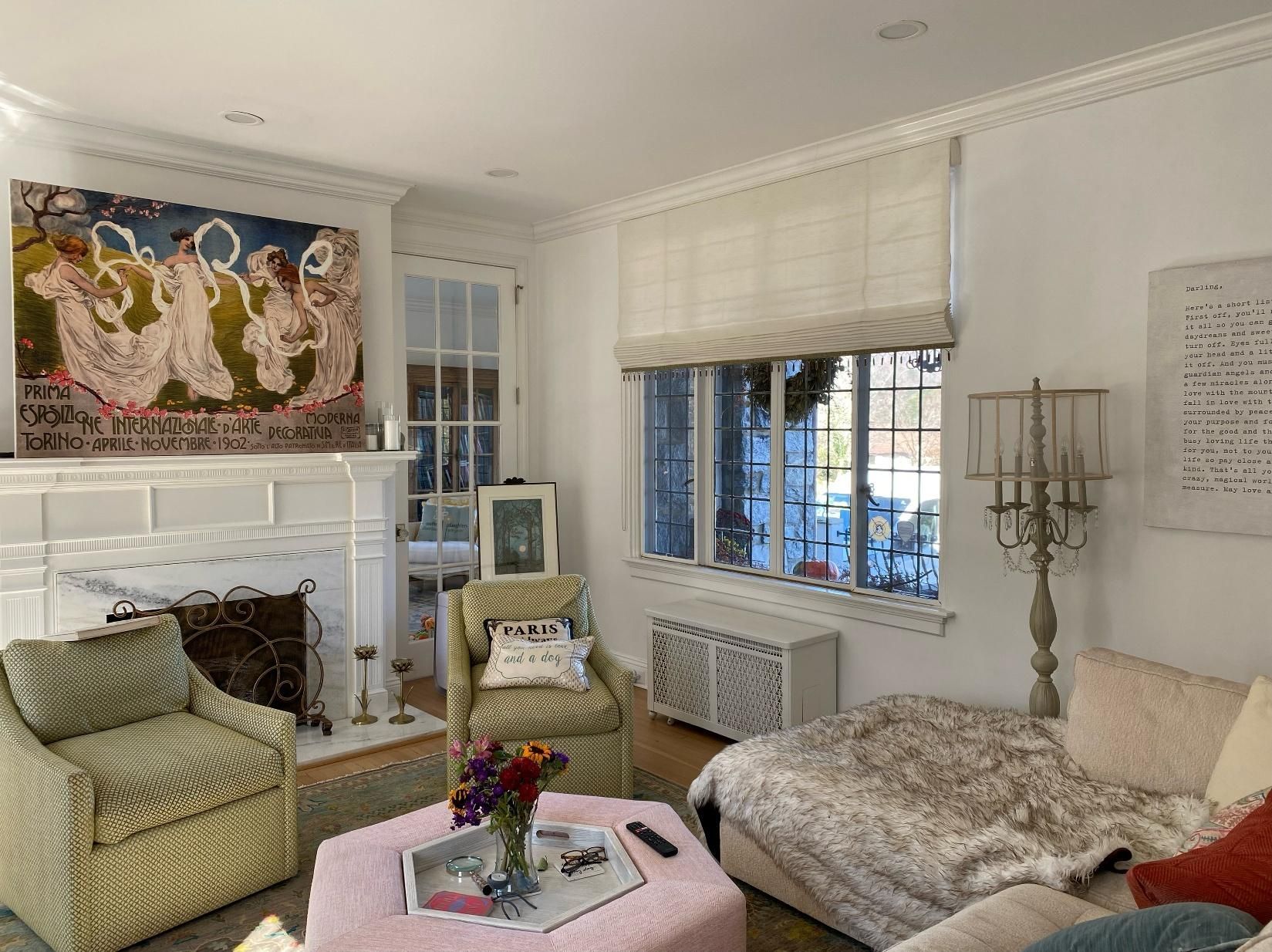Elegant living room with white walls, floral sofa, pink ottoman, fireplace, and large windows with blinds