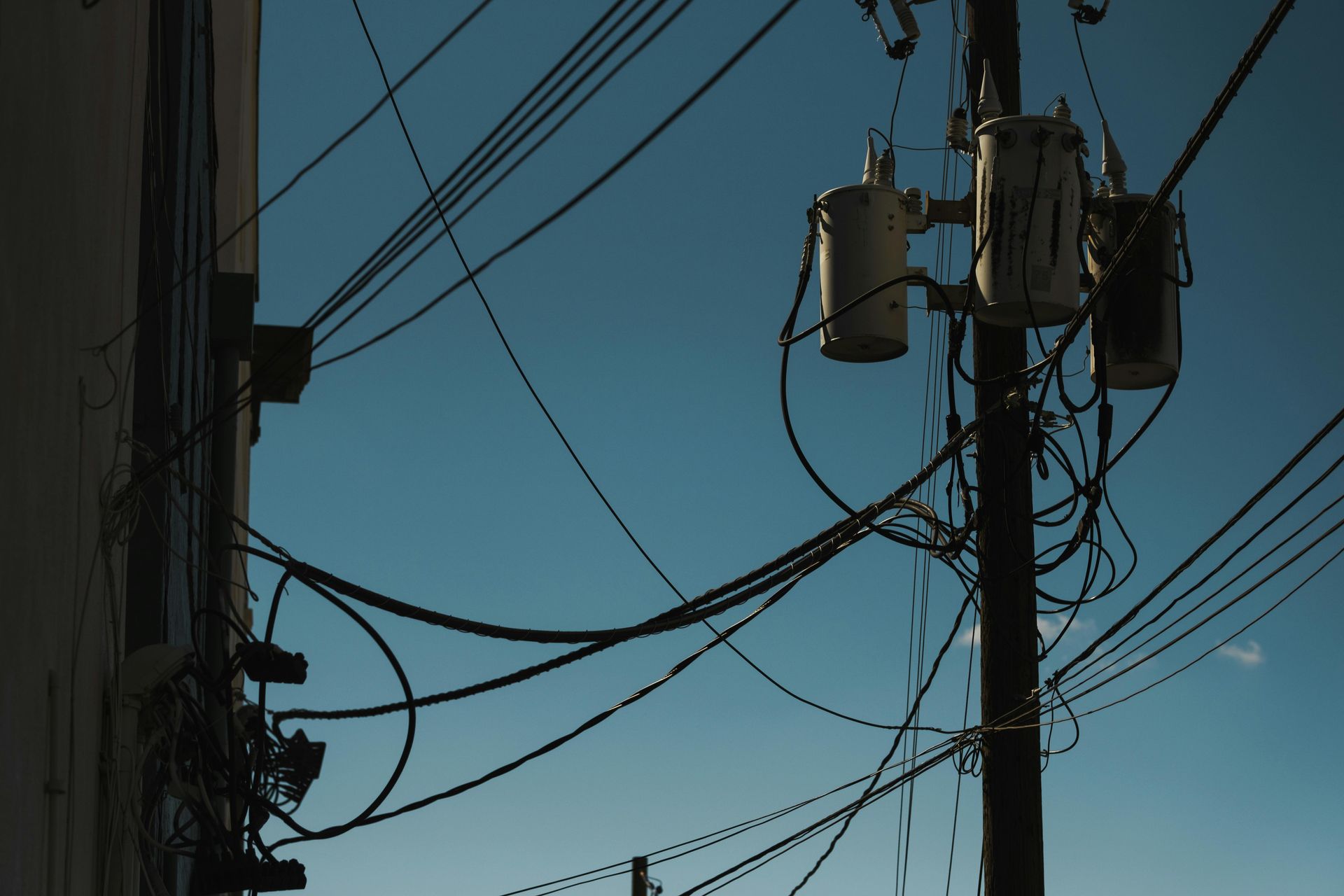 A tangle of power lines and transformers against a clear blue sky, next to a building.