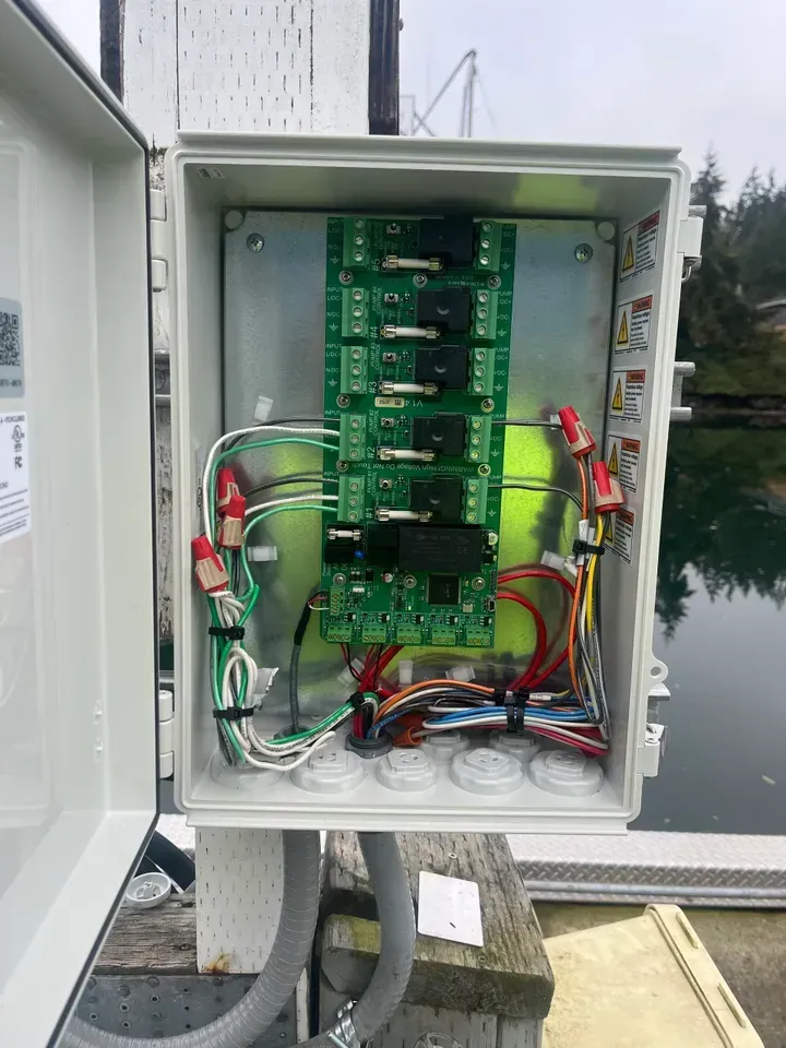 Open white electrical box on a dock with circuit boards, wires, and connectors. The background shows water and a structure.