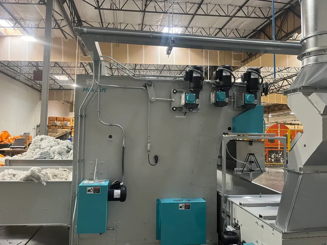 Machinery in a factory: gray and teal industrial equipment with ventilation pipes, against a neutral wall.