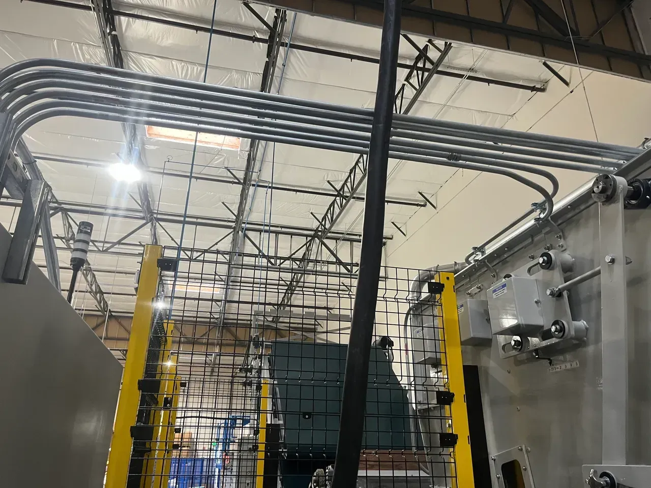 Metal pipes run across a ceiling, with a large machine visible on the right and yellow supports on the left.