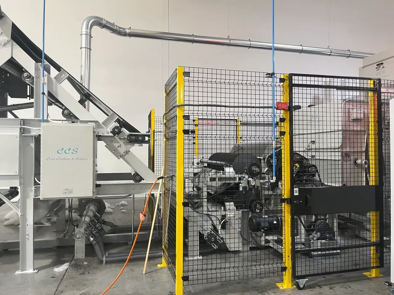Industrial machine behind yellow safety fencing. Conveyor belt at left; silver tubing overhead.