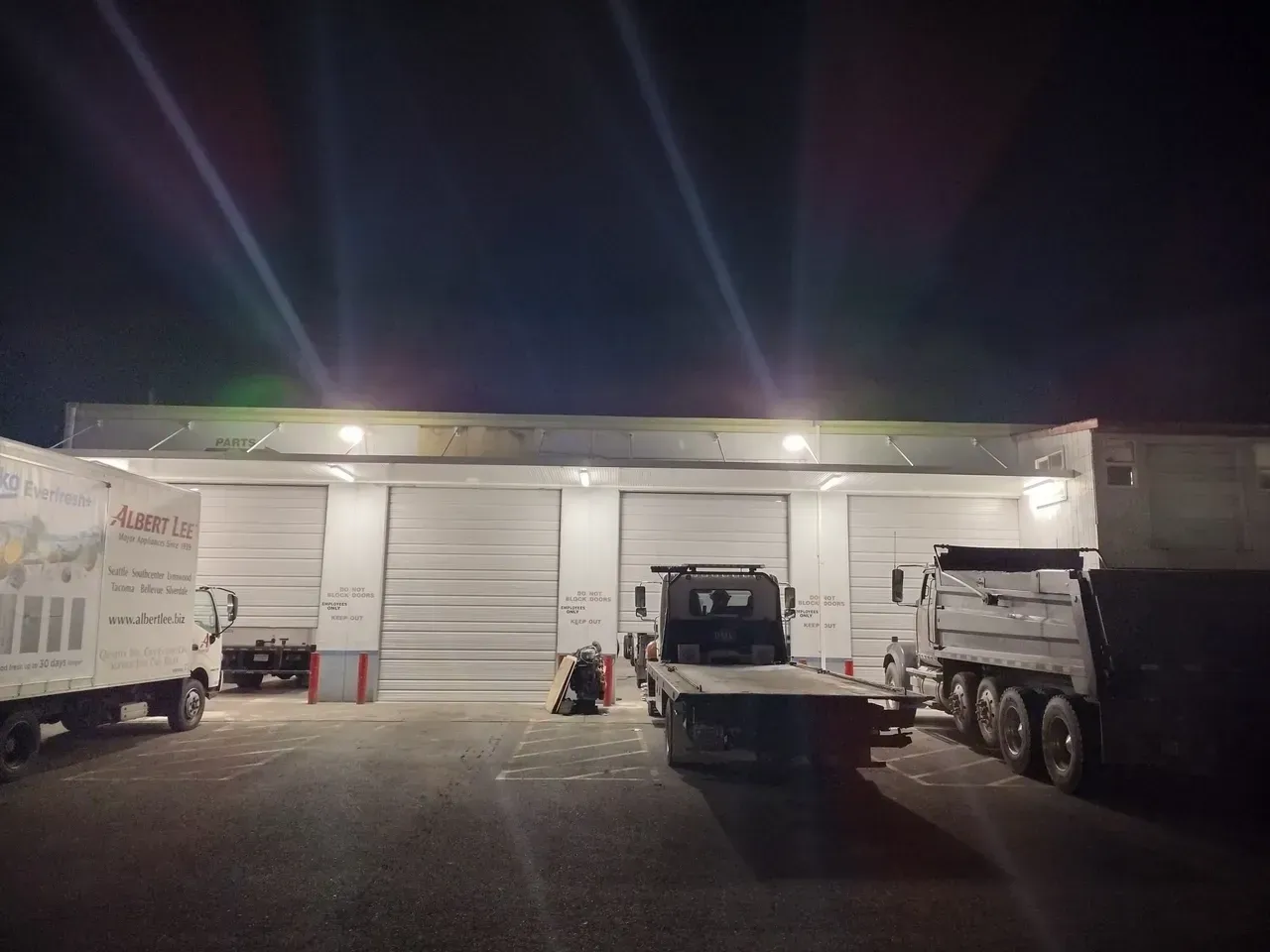 Exterior of a building with three garage doors, tow truck, and two other trucks at night.