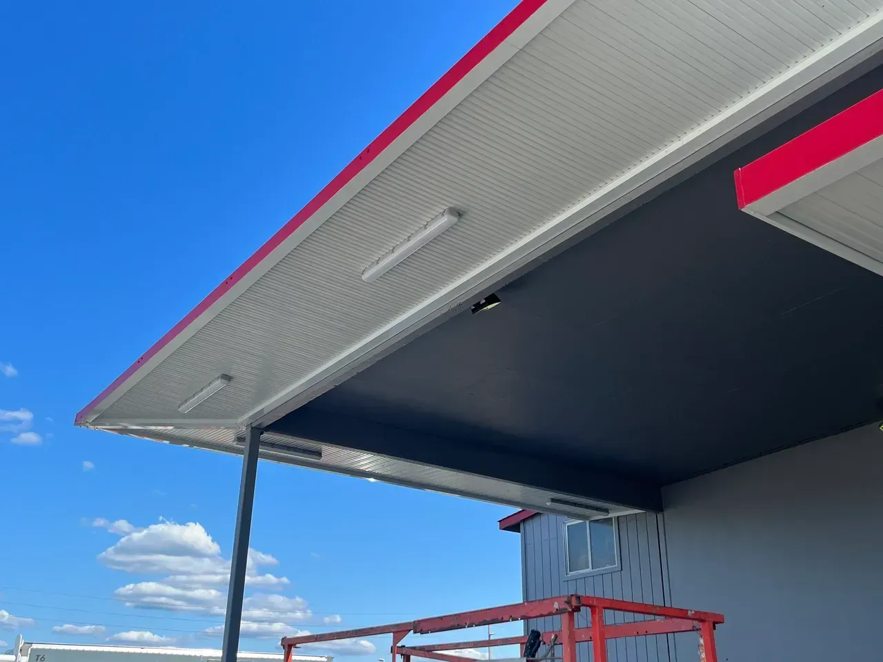View of a building's canopy with a red trim, white corrugated ceiling, and black underside against a blue sky.