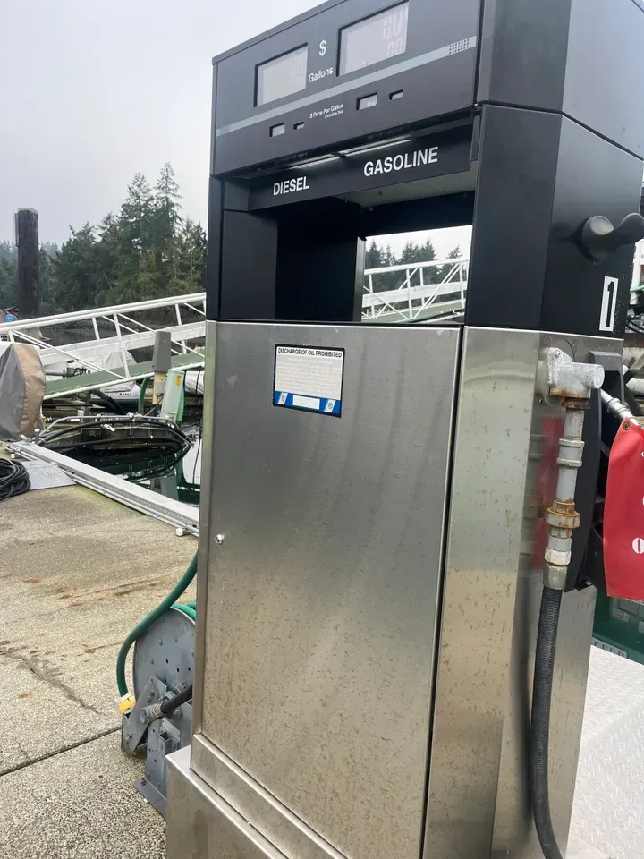 Gasoline pump on a dock. Stainless steel with digital displays. Green hose and water in the background.