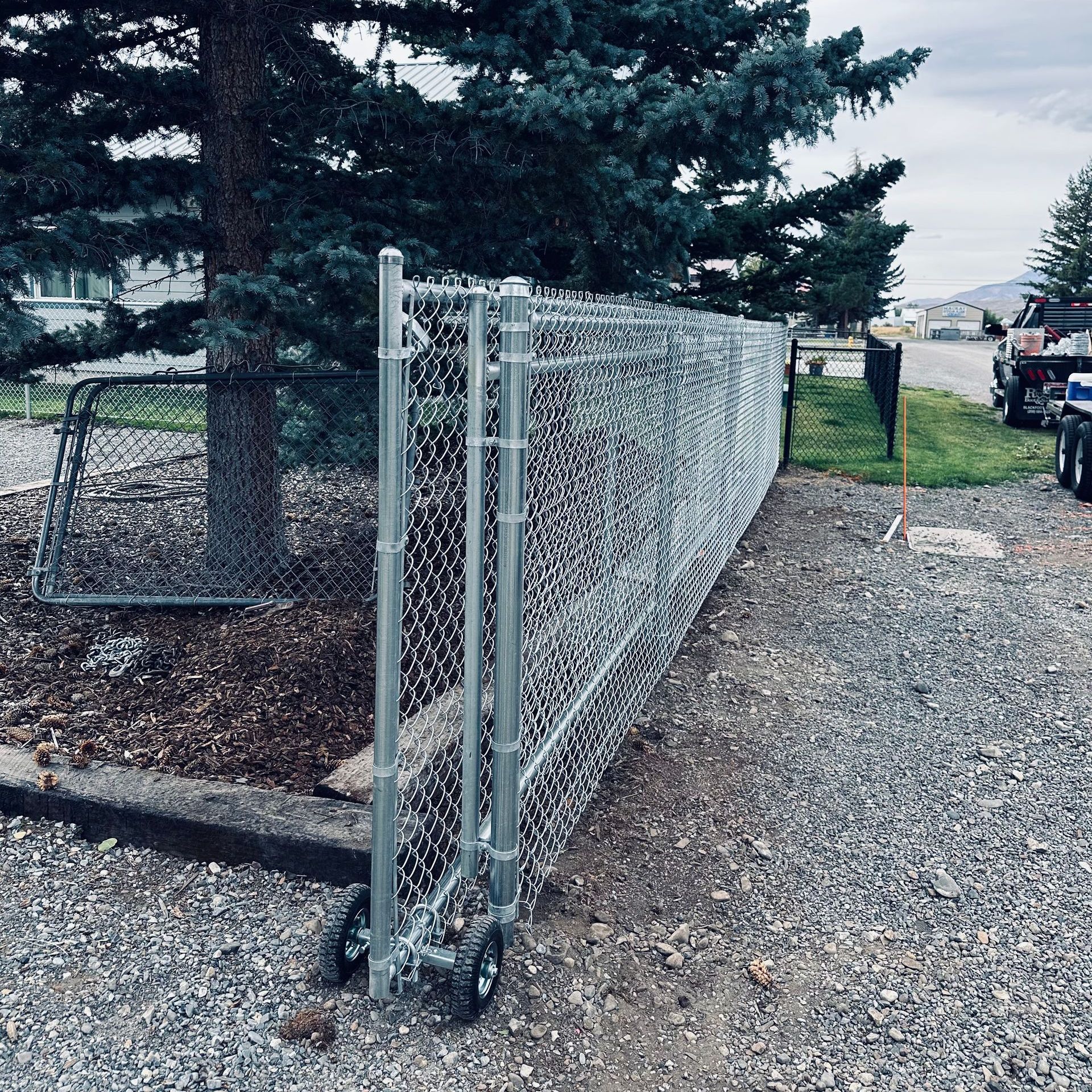 A chain link fence with wheels is sitting on gravel next to a tree.