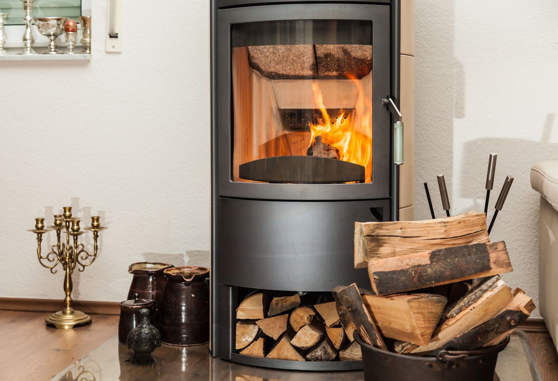 Lit wood-burning stove in a living room beside stacked logs and fireplace tools