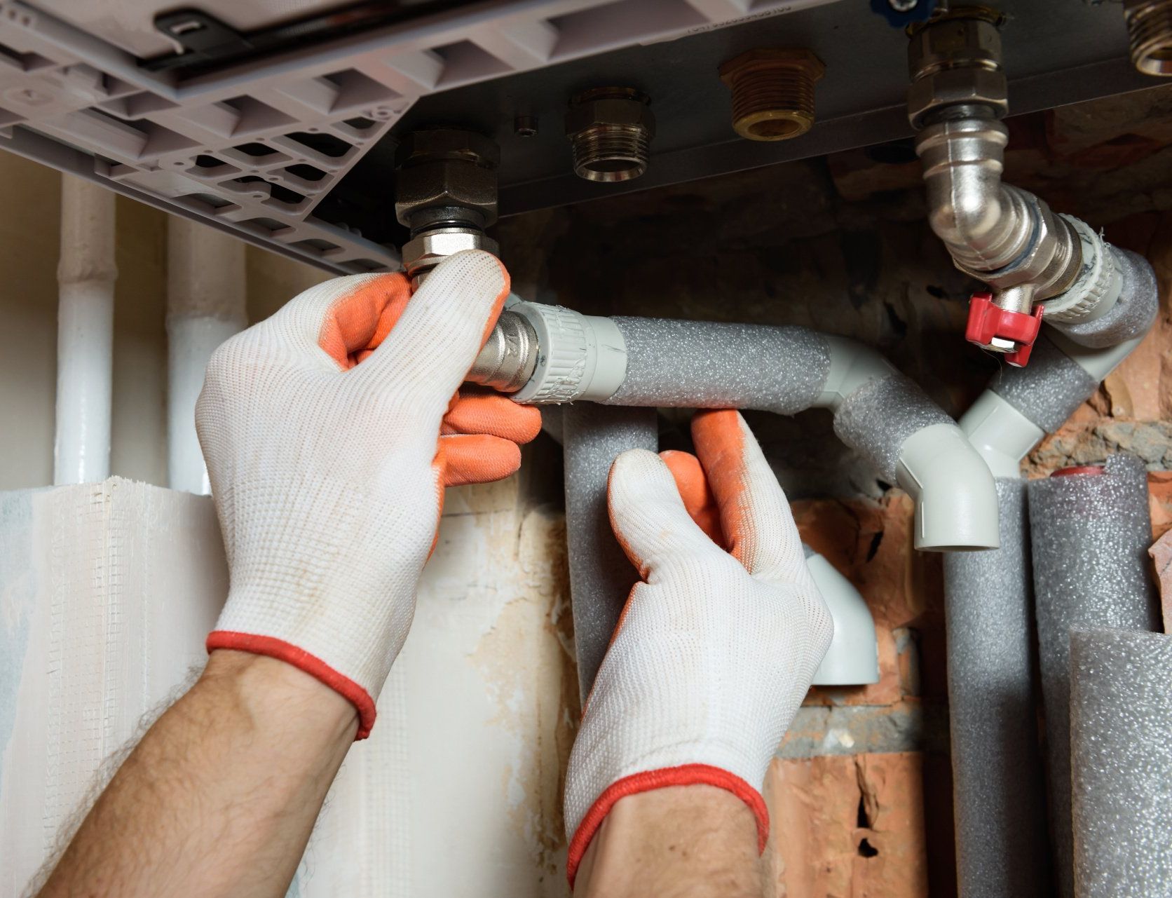 Gloved hands tightening insulation on metal pipes under a sink or appliance