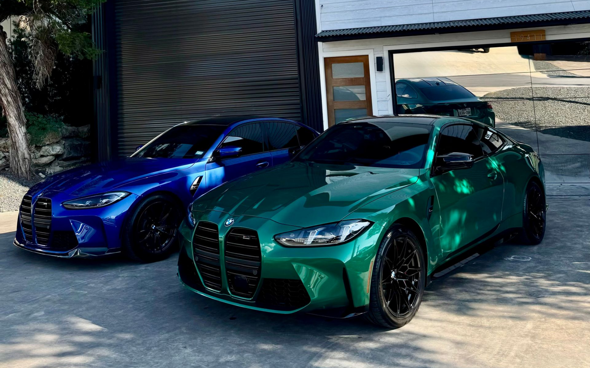 Two parked BMW M4 cars, one blue, one green, in front of a modern building with a garage.