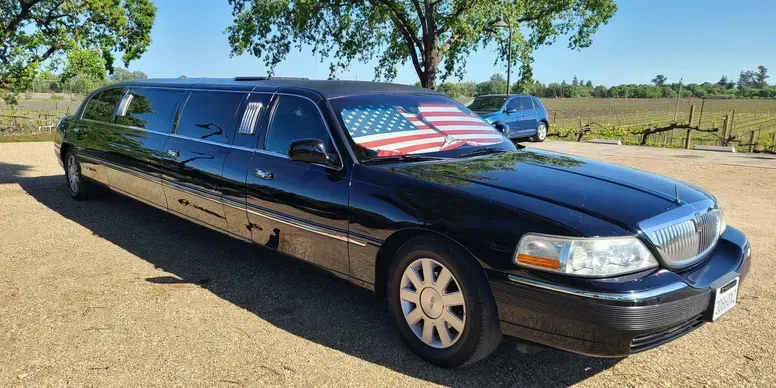 A black limousine with an american flag on the windshield is parked in a parking lot.