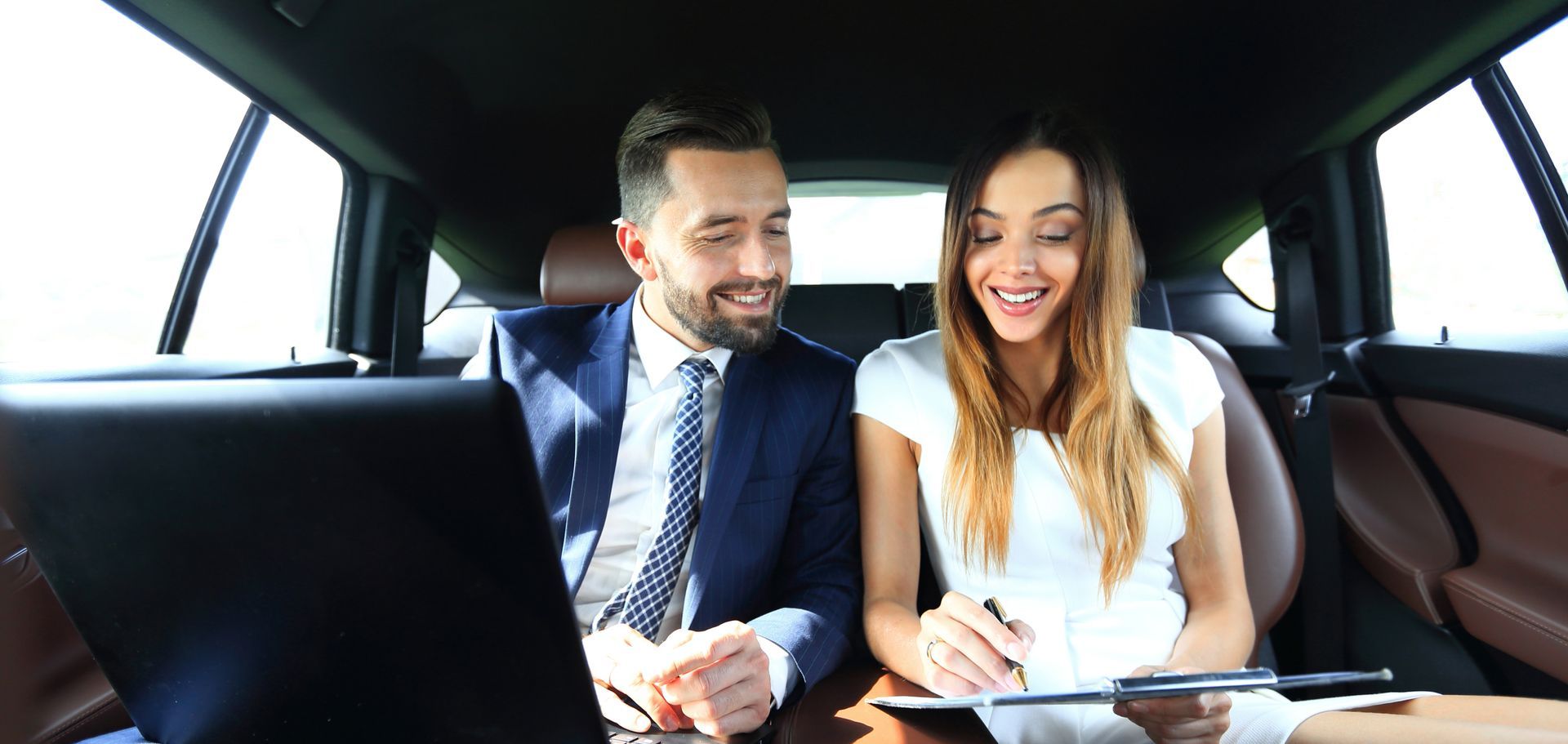 A man and a woman are sitting in the back seat of a car looking at a laptop.