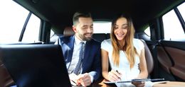 A man and a woman are sitting in the back seat of a car looking at a laptop.