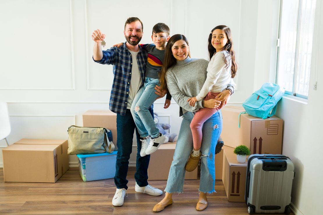 Family standing in a bright room with moving boxes, smiling and waving during a move