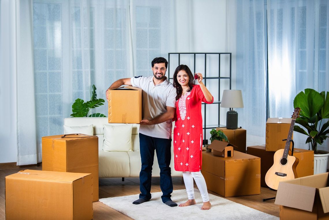 Smiling couple in a furnished room surrounded by moving boxes, holding packed items and giving thumbs up