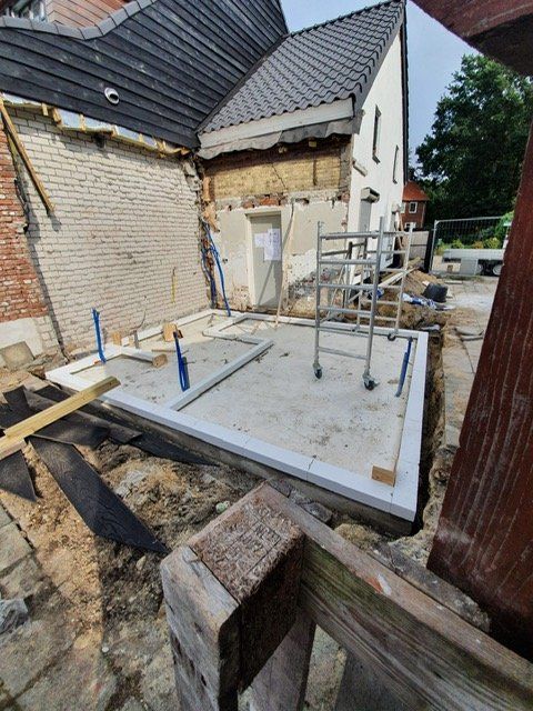 Bouwplaats: betonnen fundering, huis van baksteen en wit, steigerwerk, zichtbare leidingen.