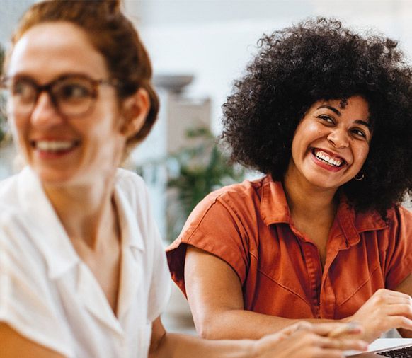 Duas mulheres estão sorrindo enquanto estão sentadas à mesa com um laptop.