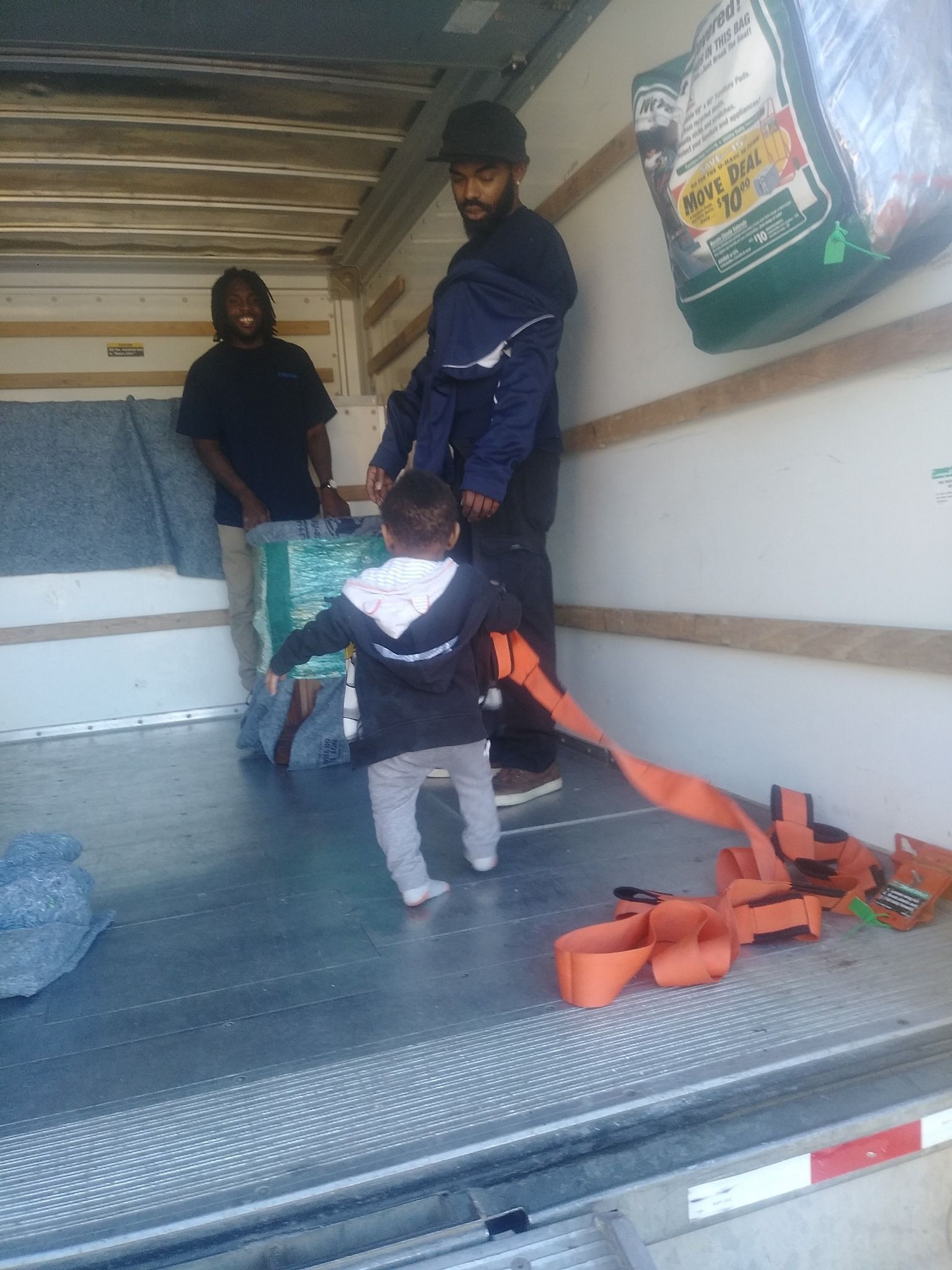 Inside moving truck: two men carrying boxes, child walking, orange straps on floor.