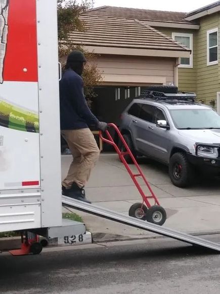 Person wearing a black mask using a hand truck to unload items from a moving truck in front of a house.