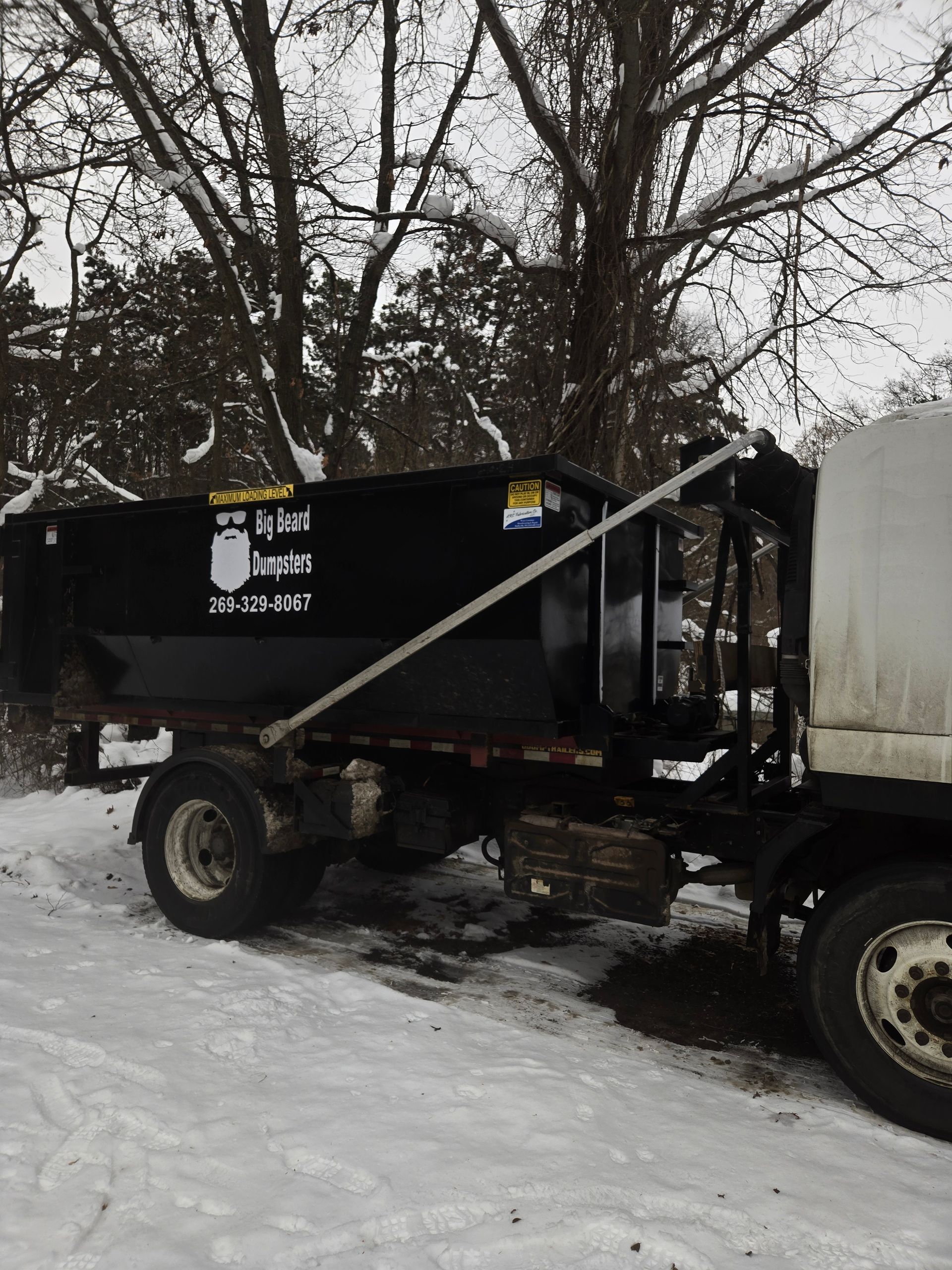 Black dumpster on a truck in a snowy area, trees in background.