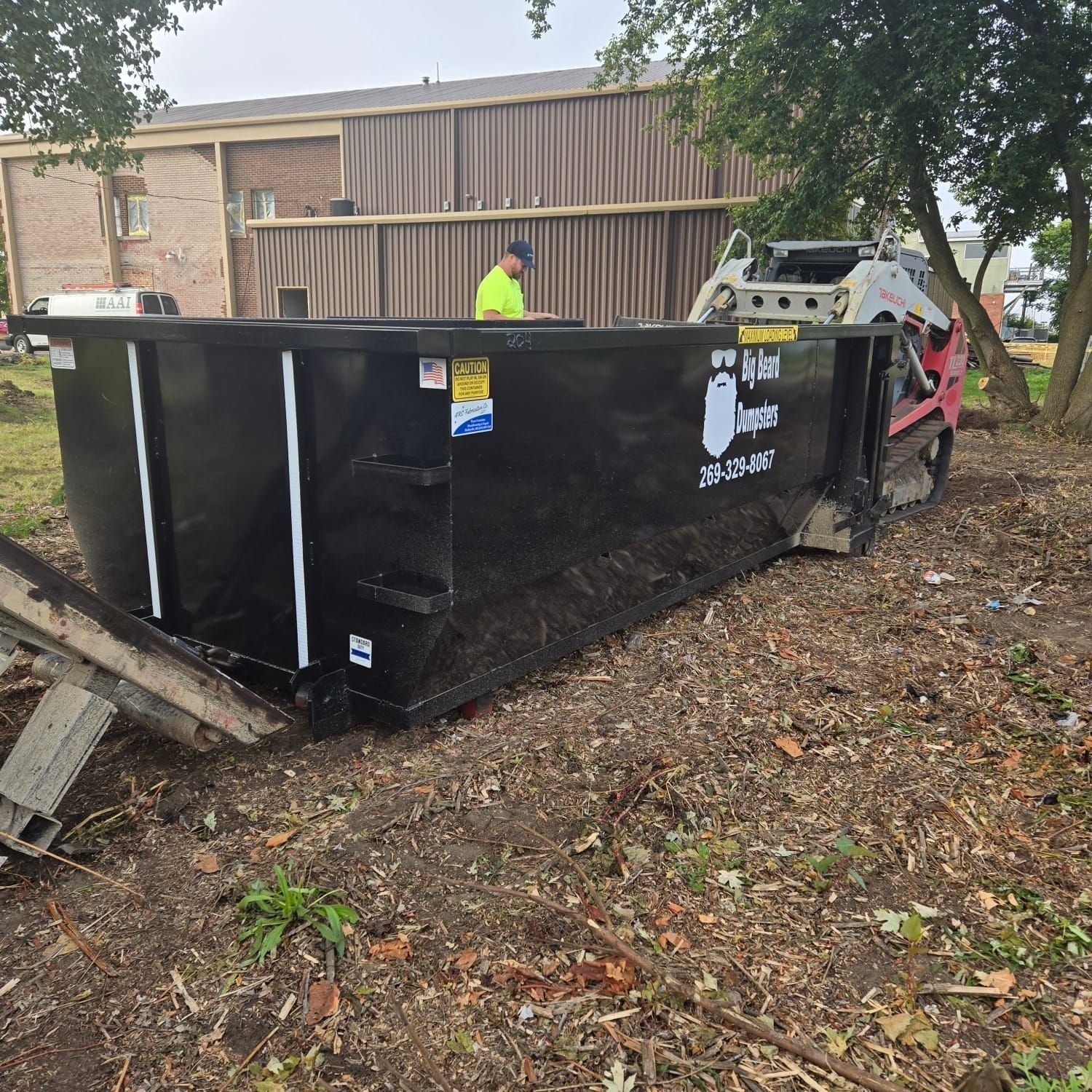 A large black dumpster is sitting in the dirt in front of a building.