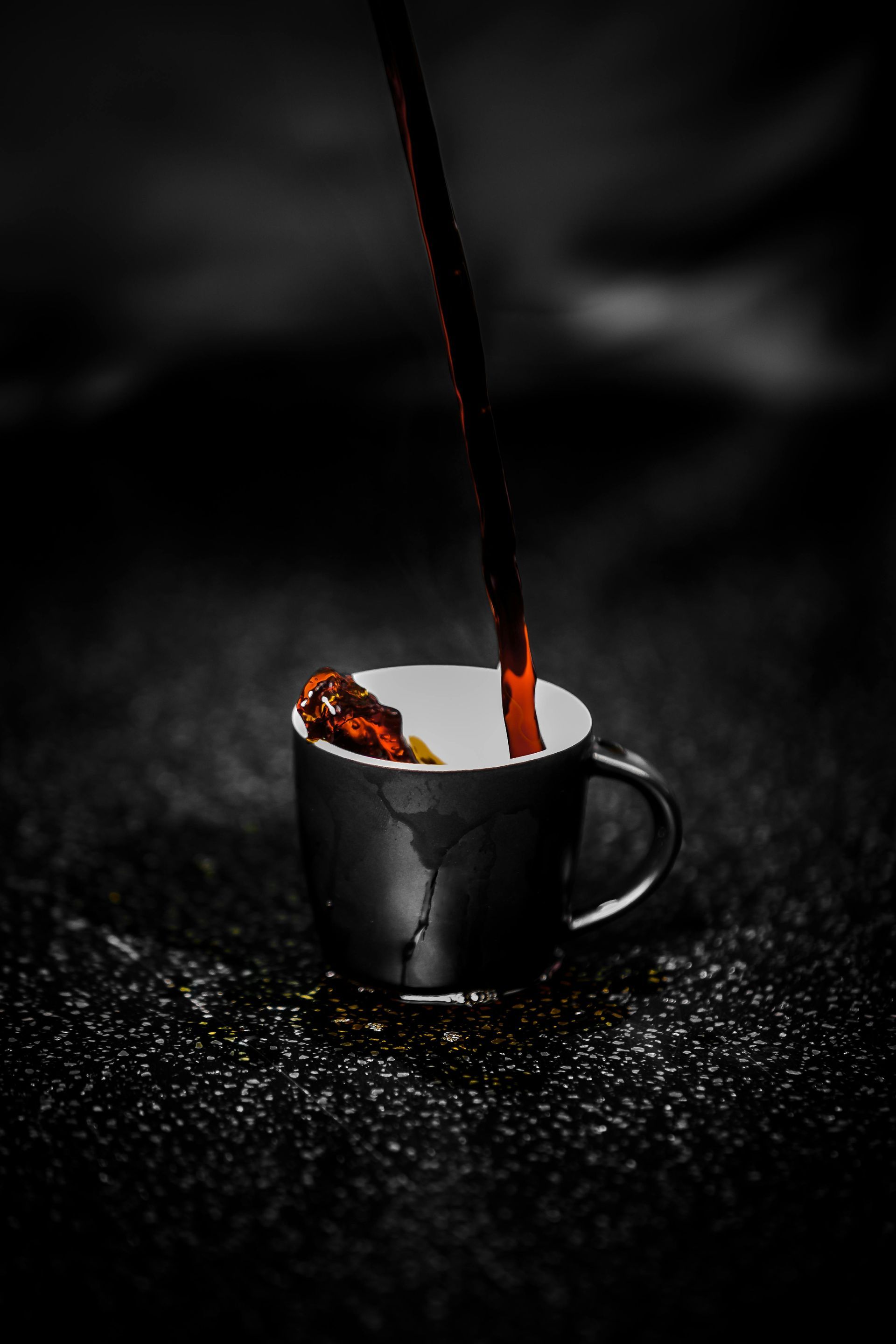 Dark liquid pouring into a black mug, with some liquid and spices visible; set on a dark surface.