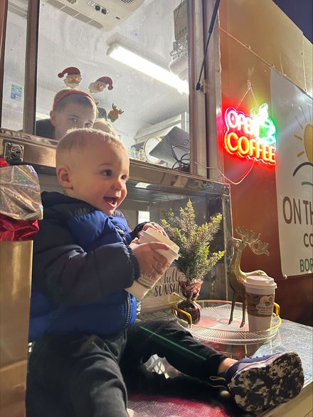 Boy in blue vest smiles, holding container near coffee shop window with