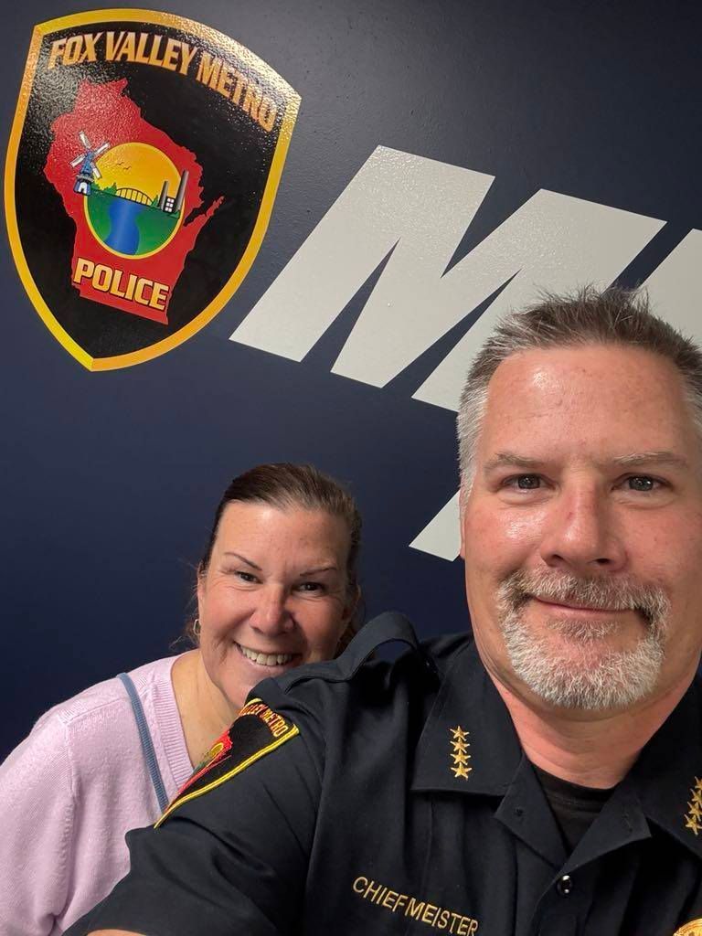 A man and a woman are posing for a picture in front of a fox valley metro police badge.