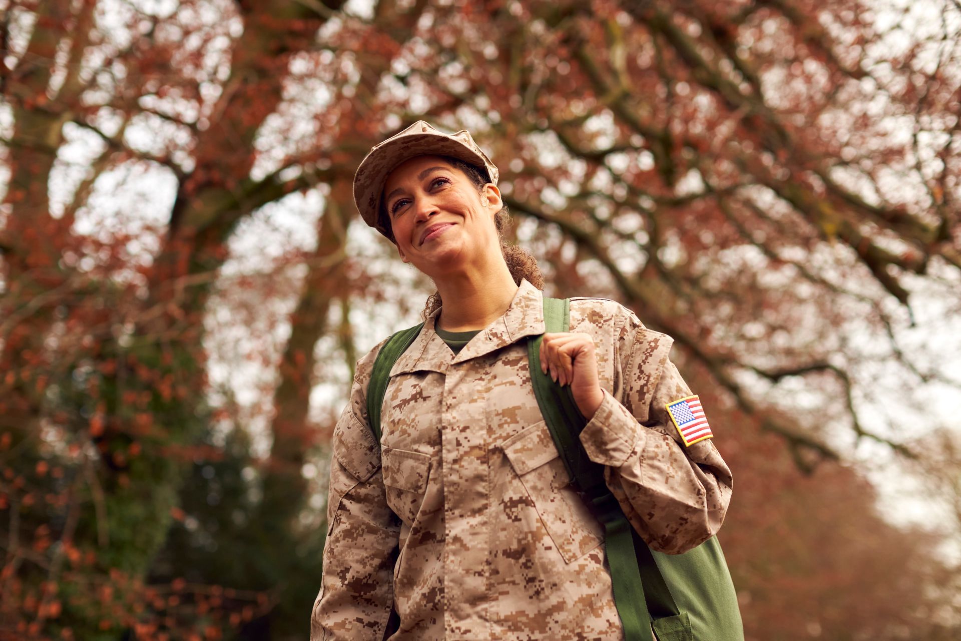 A woman in a military uniform is carrying a backpack and smiling.